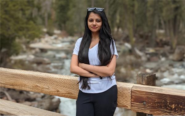 Person with long black hair standing in front of woods