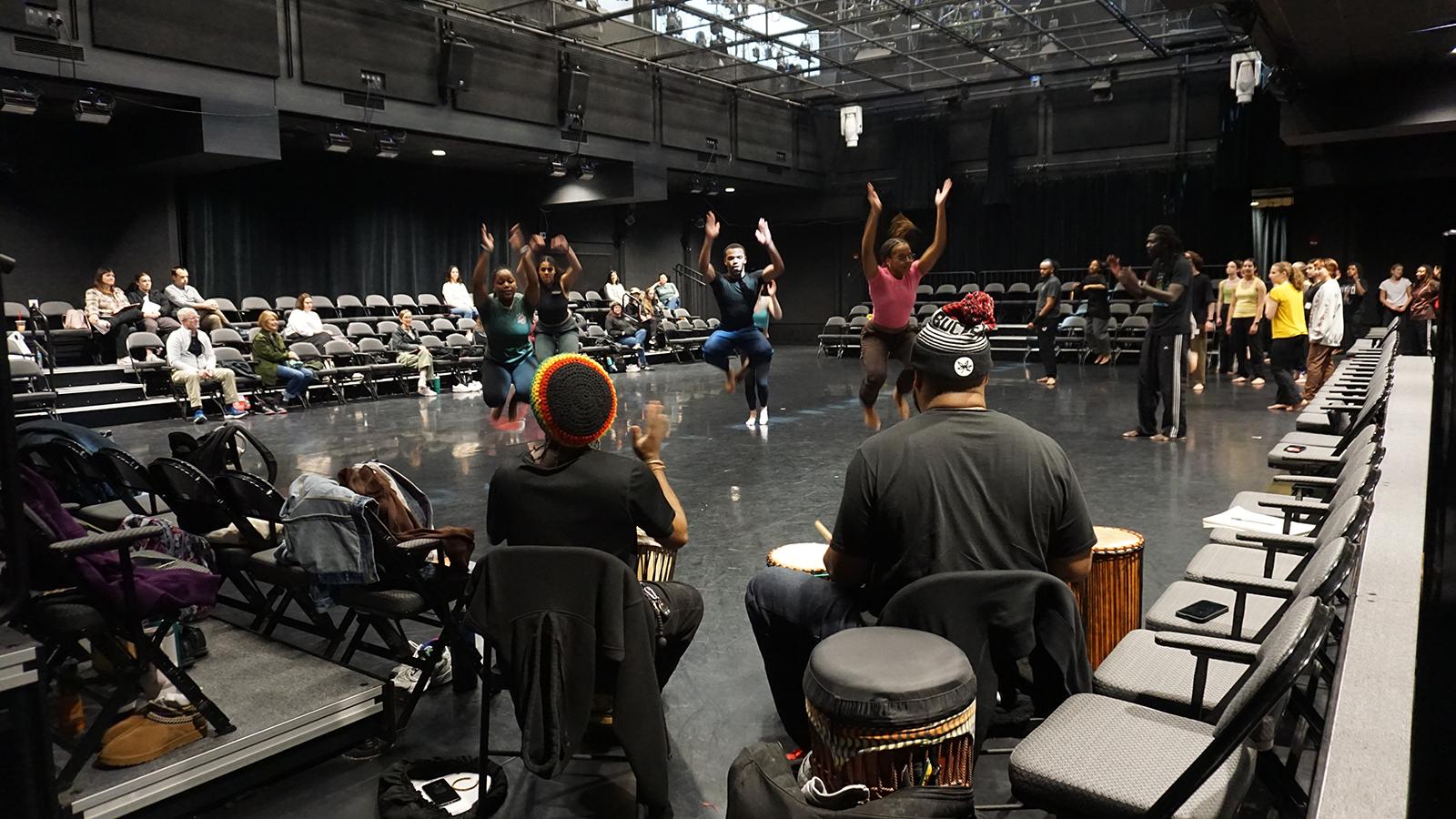 a group of dancers leaping in front of drummers in a theatre